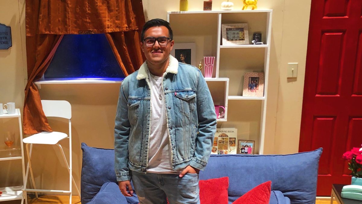 A man in blue jeans and a blue jean jacket stands in front of a set with a couch, a window with curtains, a bookshelf, and a red door.