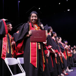 Graduates on stage with female holding her diploma cover and smiling