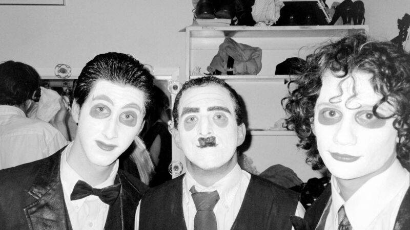 Three men wearing suits and stage makeup behind the scenes of a production