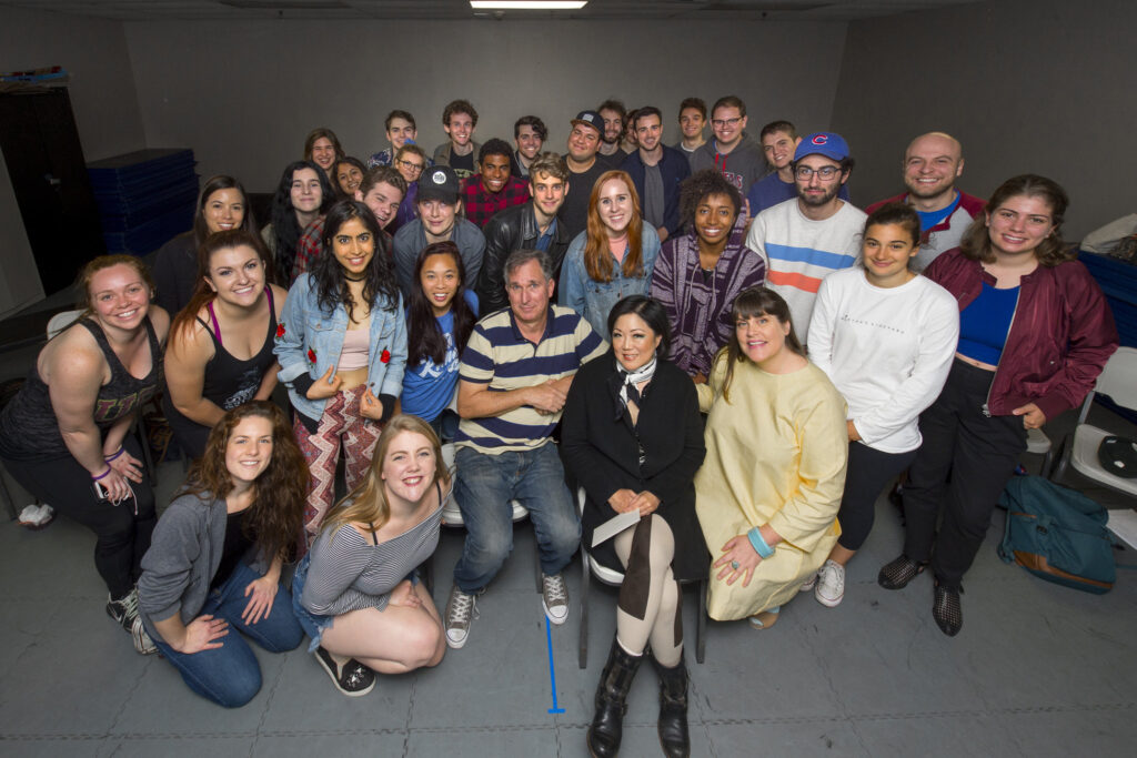 A group photo of a class of students in a grey classroom, surrounding Wayne Federman and Margaret Cho.