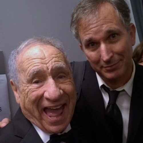 An older man on the left smiles with mouth open for a selfie next to a man smiling on the right, both dressed in suits.