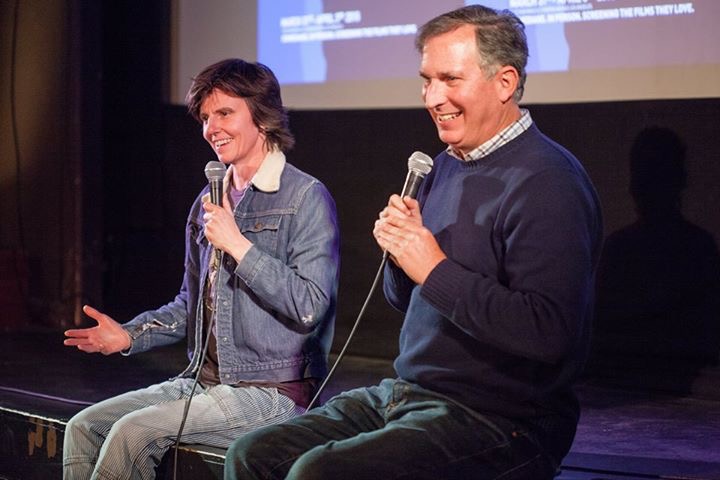 A man sits on the edge of a stage, arms folded with microphone to mouth, and a woman sits on the left, gesturing as she speaks into her microphone.