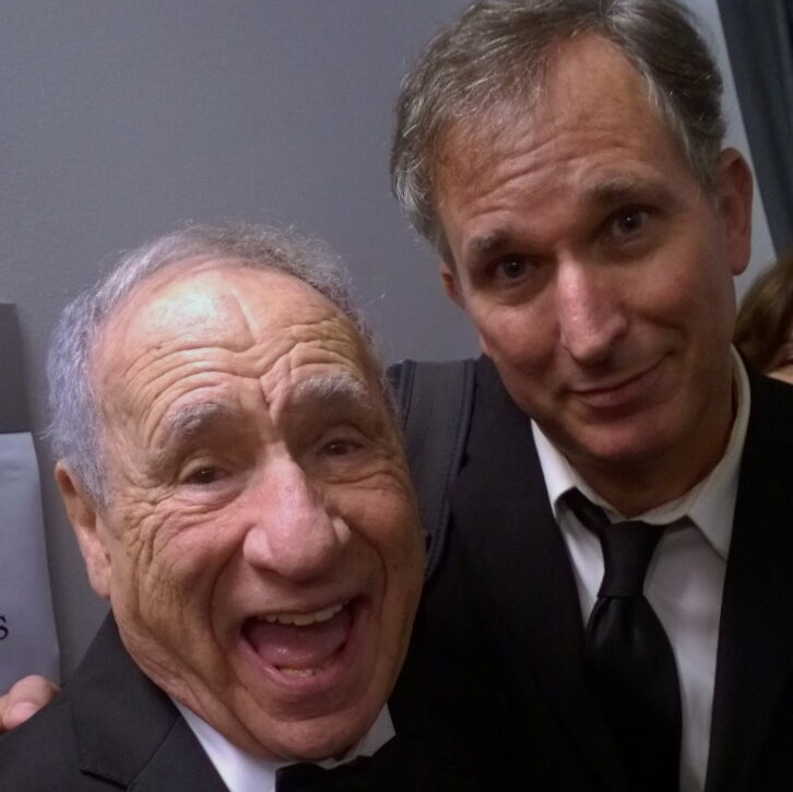 An older man on the left smiles with mouth open for a selfie next to a man smiling on the right, both dressed in suits.