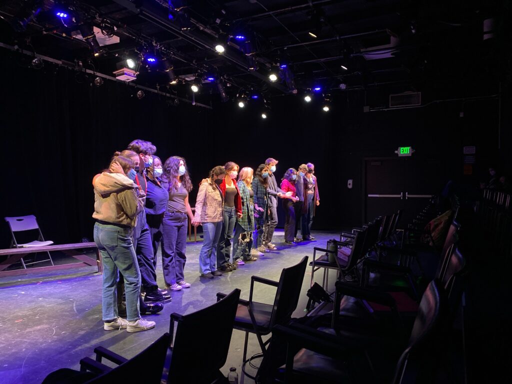 Twelve student-actors joining hands on stage in a rehearsal, wearing masks. 