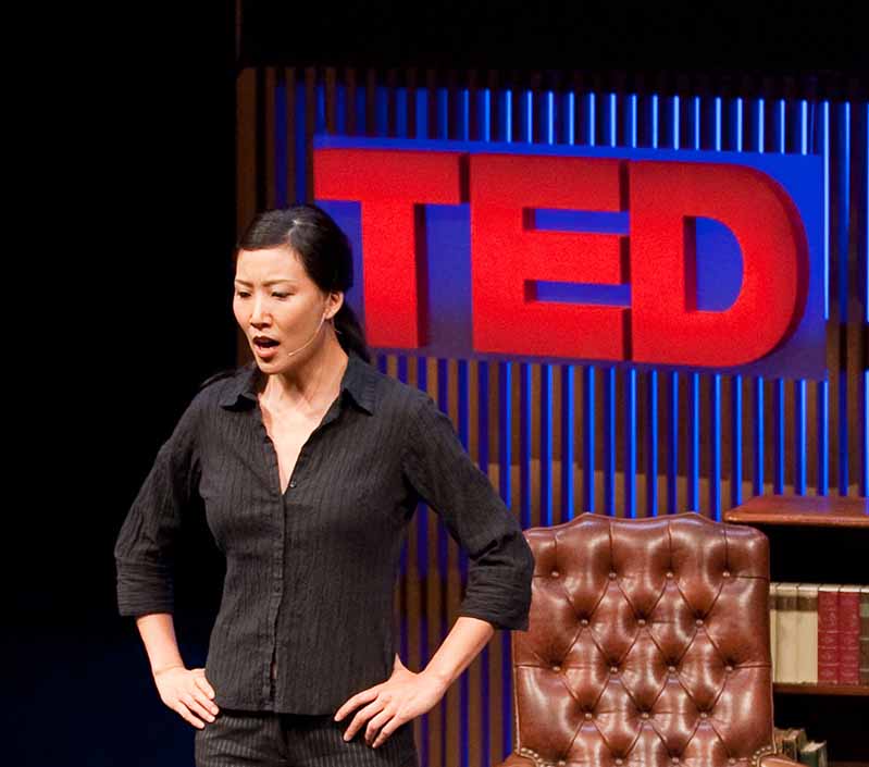 Esther K. Chae stands with hands on hips in front of a red TED sign, performing a monologue.