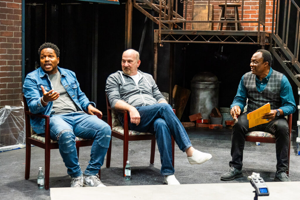 (l to r): Sheldon Candis, Rob Schiller and Bayo Akinfemi sit side by side on chairs. Akinfemi and Schiller listen as Candis gestures.