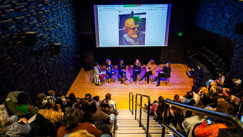 Six artists sit onstage discussing a projected image of a 