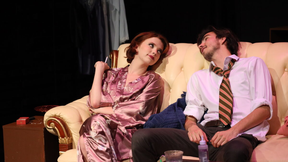 A female and male actor in costumes sitting on a couch as part of a performance of Dry Land.