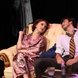 A female and male actor in costumes sitting on a couch as part of a performance of Dry Land.