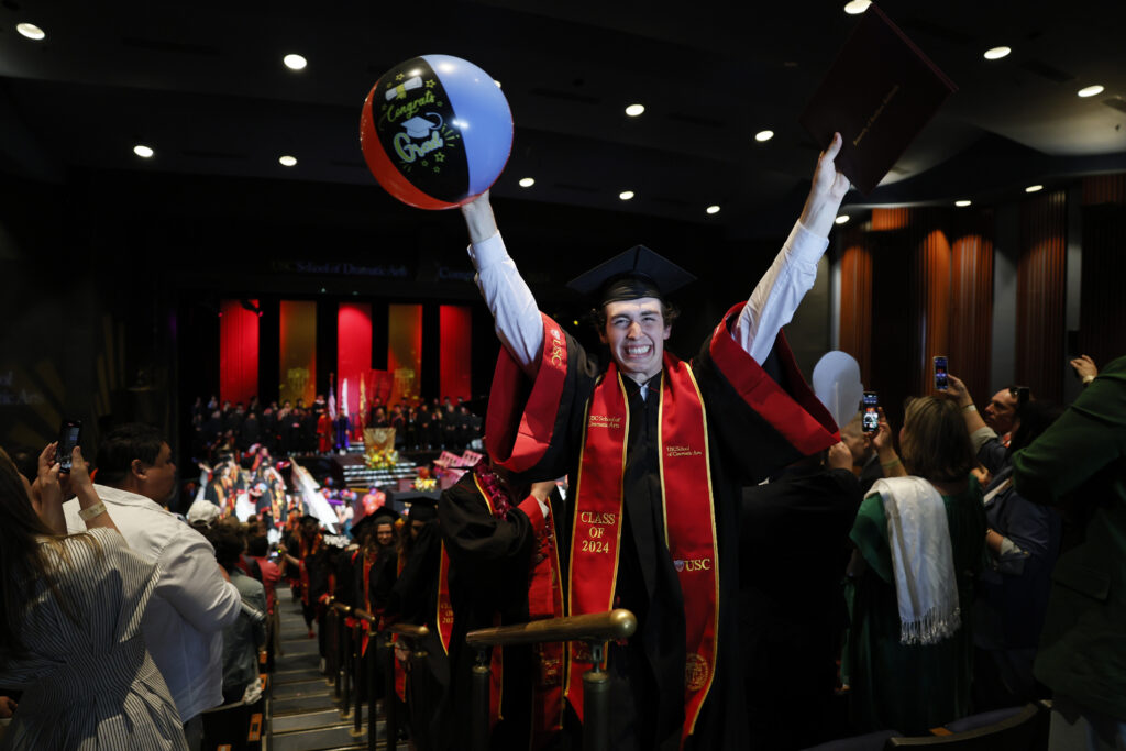 A graduating student raises both arms and holds a beachball in celebration.