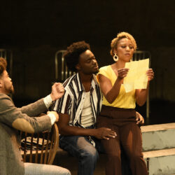 The cast of King Hedley the second perform on stage, with Mister gesturing toward King Hedley and Tonya. Tonya is reading a note while King watches on.