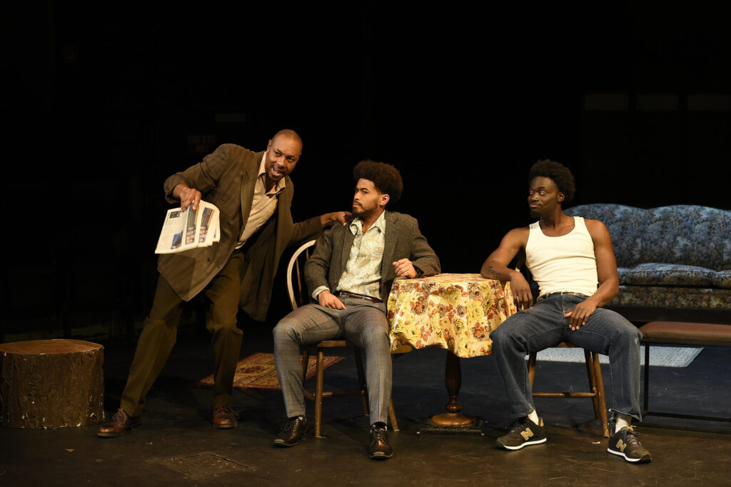 Stool Pigeon, Mister and King Hedley sit side by side. Stool Pigeon is holding a newspaper and leaning in to the group to tell a story.