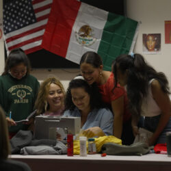 A group of actors huddling around a sewing machine with the Mexican and American flags in the background.