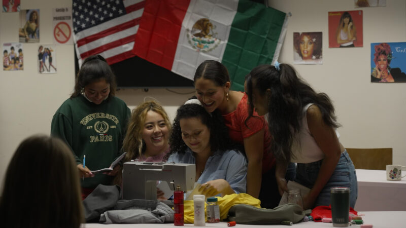 A group of actors huddling around a sewing machine with the Mexican and American flags in the background.