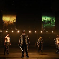 Four actors stand on a lighted stage, the actor in the foreground holding a sword.