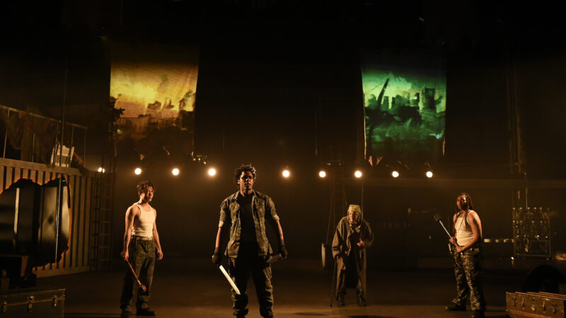 Four actors stand on a lighted stage, the actor in the foreground holding a sword.