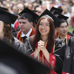 A student in commencement regalia clasps her hands together and looks toward the stage with joy.