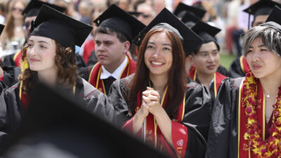 A student in commencement regalia clasps her hands together and looks toward the stage with joy.
