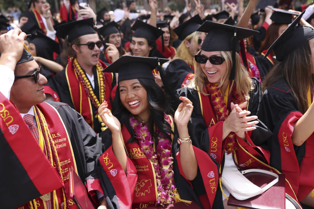 Graduates in commencement regalia clap and laugh as they celebrate with one another.
