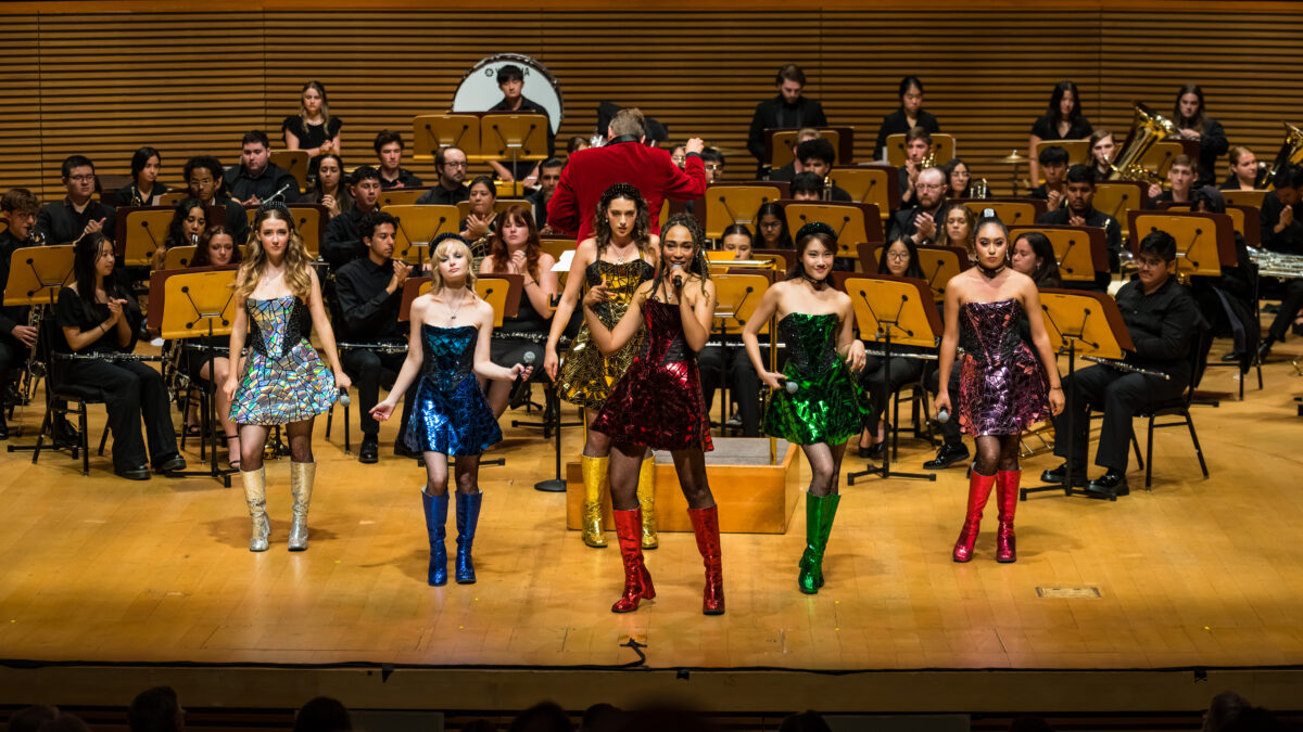 Photo of six women in colorful costumes in front of an orchestra