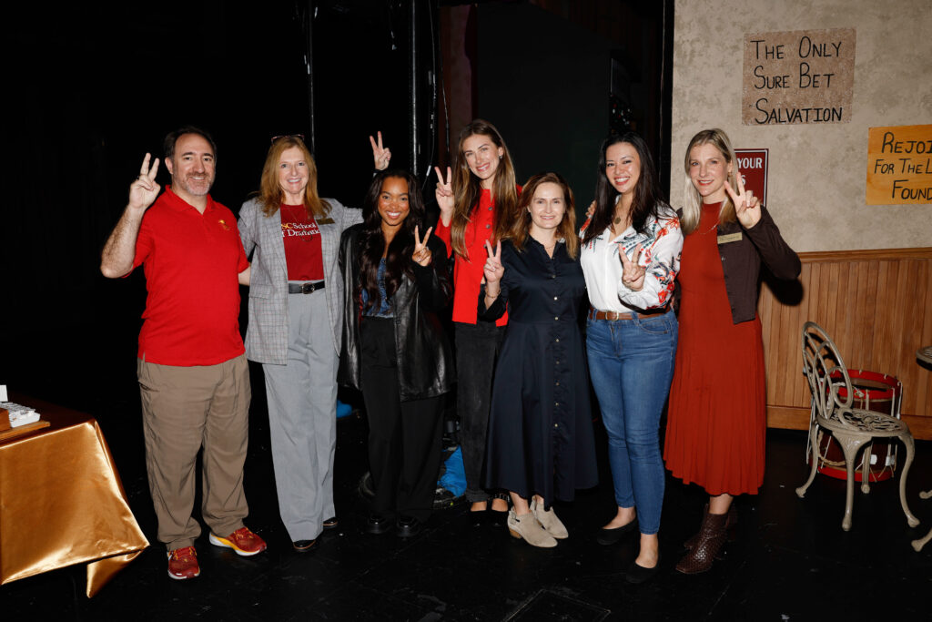 SDA alumni panelists, moderator Debi Manwiller and Dean Emily Roxworthy pose together backstage, flashing victory signs, with theatrical set pieces visible in the background.