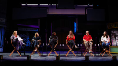 Six panelists seated in director's chairs on stage in the Bing Theatre during the SDA alumni panel at Trojan Family Weekend, with a theatrical set backdrop behind them