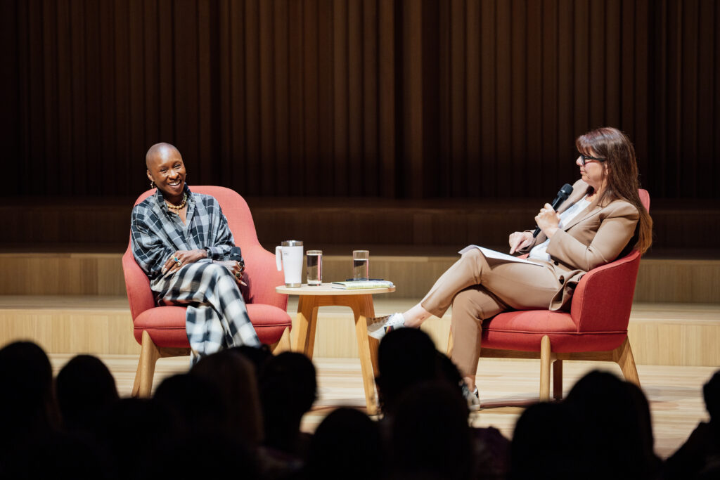 Cynthia Erivo, seated in a red armchair on stage, smiles during a conversation with Dr. Stacy L. Smith, who is seated across from her holding a microphone and notes. A small round table with water glasses sits between them. The silhouettes of a full audience are visible in the foreground, with warm wood paneling behind the stage.