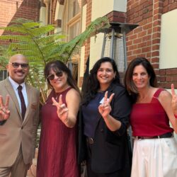 Four people stand together outside a brick campus building, smiling and making the Trojan 