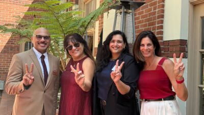 Four people stand together outside a brick campus building, smiling and making the Trojan 